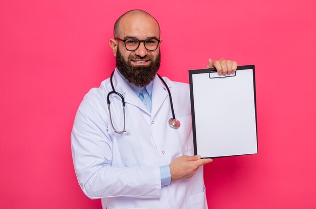 Feliz barbudo médico com jaleco branco com estetoscópio em volta do pescoço usando óculos segurando uma prancheta com páginas em branco olhando com um sorriso no rosto