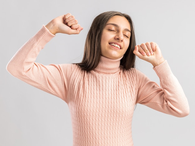 Feliz bastante adolescente manteniendo los puños arriba con los ojos cerrados aislado en la pared blanca