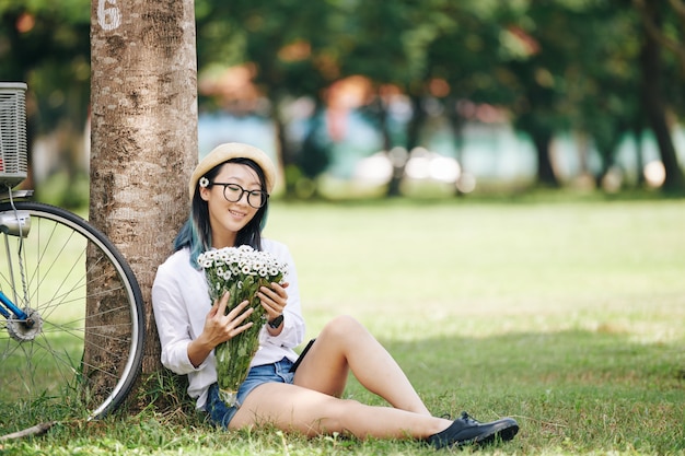 Feliz bastante joven China sentada bajo un árbol en el parque y loonig en ramo de margaritas en sus manos