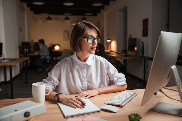 Feliz bastante joven empresaria en gafas escribiendo y usando la computadora en la oficina