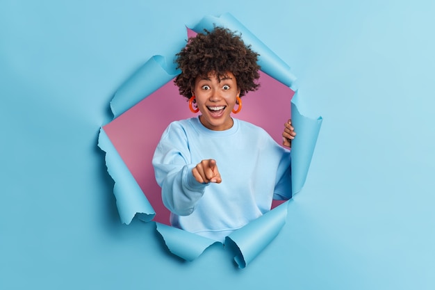 Feliz bastante joven mujer afroamericana te señala con el dedo índice ve algo divertido en el frente viste un jersey de manga larga se rompe a través de la pared de papel