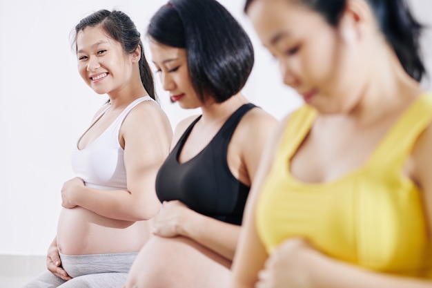 Feliz bastante joven mujer vietnamita tocando el vientre asistiendo a clases de deportes con otras mujeres embarazadas