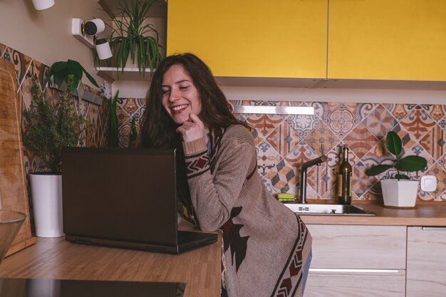 Feliz bella mujer joven en ropa casual trabajando en una computadora portátil en la cocina