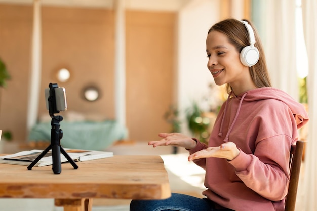 Feliz blogger adolescente famoso en auriculares inalámbricos grabando video para blog hablando con teléfono celular en trípode