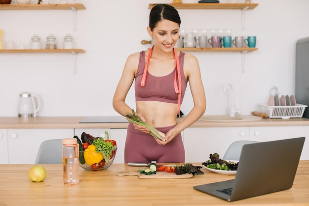 Feliz bloguera deportiva muestra espárragos y dirige una videoconferencia sobre alimentación saludable en una laptop en la cocina