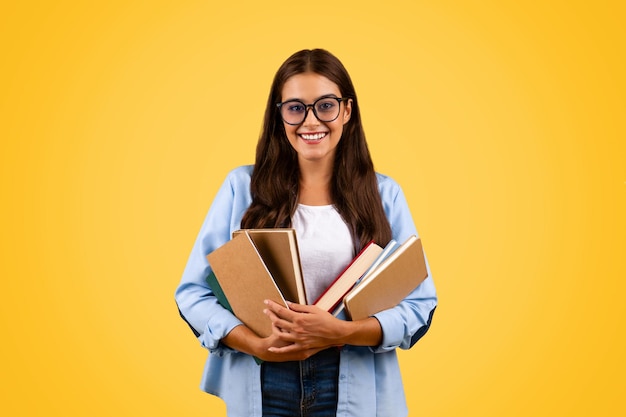 Feliz y bonita joven estudiante con gafas sostiene muchos libros aislados en el fondo amarillo del estudio