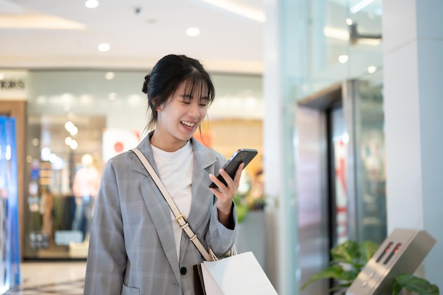 Una feliz y bonita mujer asiática está revisando mensajes en su teléfono mientras camina por un centro comercial