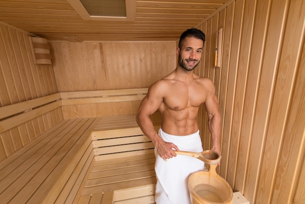 Feliz, bonito e atraente jovem com corpo musculoso, derramando água em rochas quentes na sauna