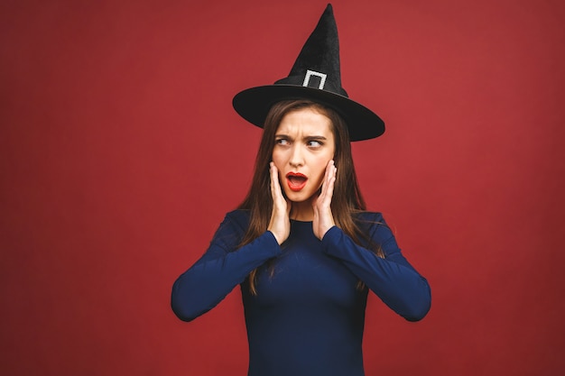 Feliz bruja de Halloween con maquillaje brillante y cabello largo. Hermosa joven sorprendida posando en traje de brujas sexy. Aislado sobre fondo rojo