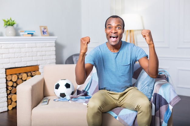 Feliz buen hombre positivo sentado en el sofá y sentirse emocionado mientras ve un partido de fútbol