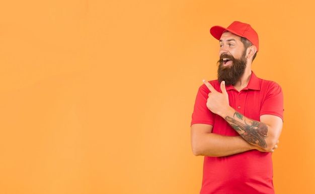 Feliz cara barbudo de boné vermelho casual e camiseta apontando o dedo para o lado do anúncio de espaço de cópia