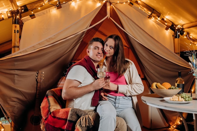 Feliz casal adorável relaxando em glamping na noite de verão e bebendo vinho perto da fogueira aconchegante Barraca de acampamento de luxo para recreação ao ar livre e recreação Conceito de estilo de vida