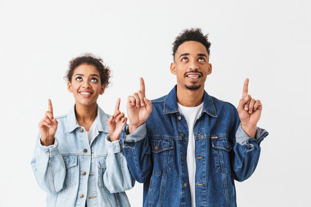 Feliz casal Africano em camisas jeans em pé juntos, apontando e olhando para cima sobre parede cinza