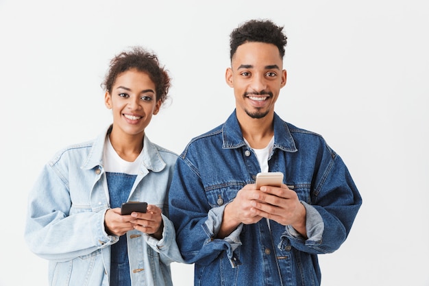 Feliz casal Africano em camisas jeans, segurando seus smartphones sobre parede cinza