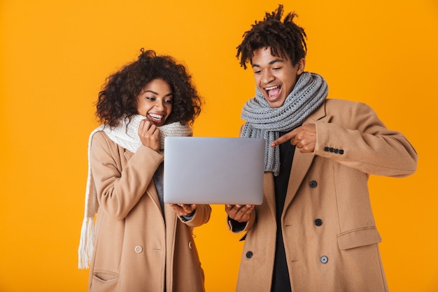 Feliz casal africano vestindo roupas de inverno, isolado, segurando um laptop, comemorando o sucesso