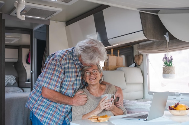 Feliz casal aposentado abraçando desfrutando de viagens de liberdade e estilo de vida alternativo viajando em caravana Mulher sênior navegando no laptop enquanto o marido a beija