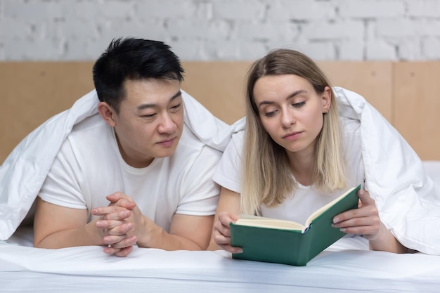 Feliz casal asiático apaixonado e mulher deitada na cama e lendo um livro