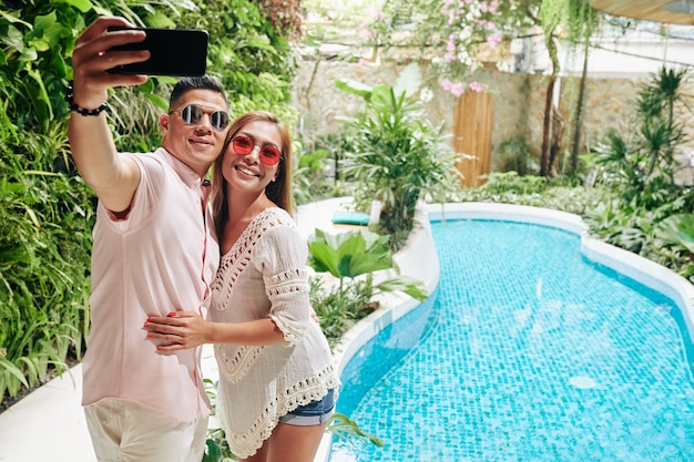 Feliz casal asiático de óculos escuros se abraçando e tirando uma selfie na piscina do spa resort