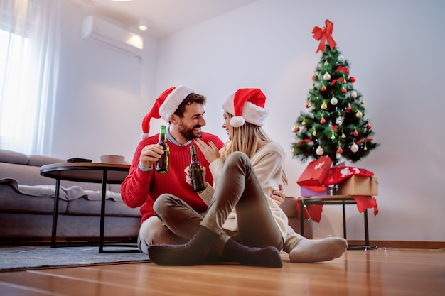 Feliz casal caucasiano bonito com chapéus de Papai Noel na cabeça, sentado no chão com uma garrafa de cerveja nas mãos e carinhos. No fundo é a árvore de Natal com presentes debaixo dela. Sala interior.