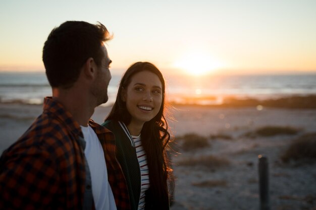 Foto feliz casal caucasiano na praia olhando um para o outro durante o pôr do sol. tempo de lazer saudável ao ar livre à beira-mar.