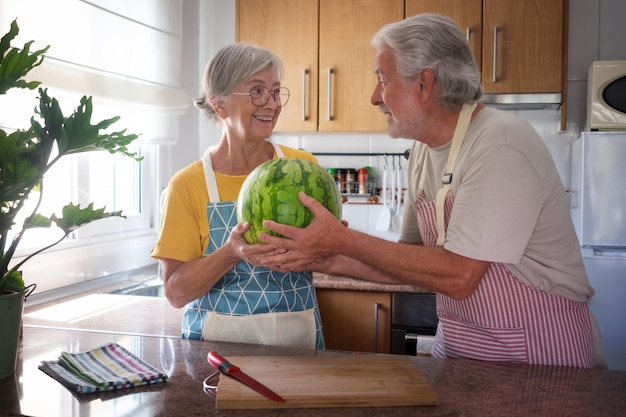 Feliz casal caucasiano sênior na cozinha de casa segurando uma melancia sazonal grande e pesada