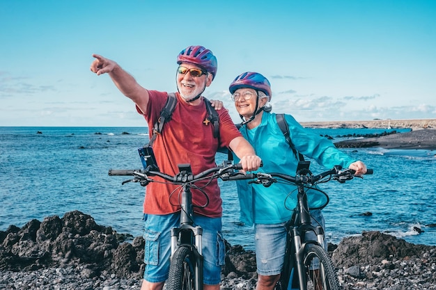 Feliz casal caucasiano sênior usando capacetes andando na praia com bicicletas elétricas Autêntico conceito de vida idosa aposentada Horizonte sobre o mar