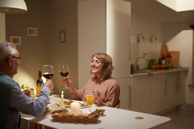 Feliz casal de idosos sentado à mesa com taças de vinho, eles jantam na cozinha de casa