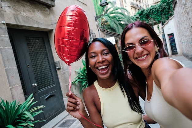 Foto feliz casal de lésbicas com um balão vermelho em forma de coração enquanto tirava uma selfie juntos ao ar livre na rua. conceito lgbt.