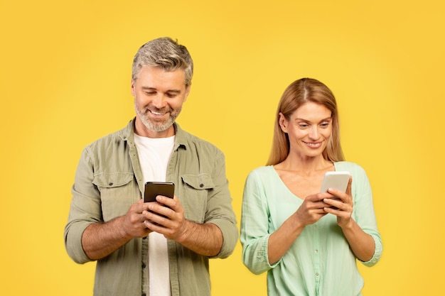 Foto feliz casal de meia-idade usando celulares e sorrindo, enviando mensagens de texto ou navegando na internet em pé no amarelo