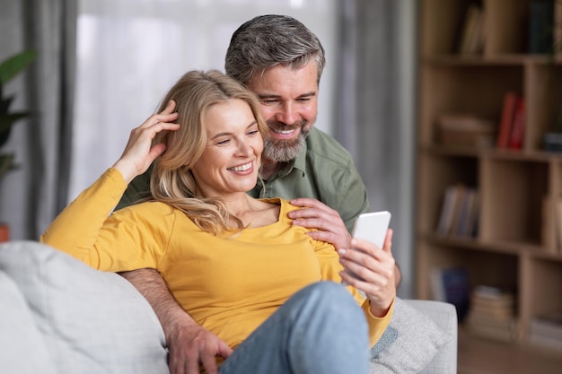 Feliz casal de meia idade usando smartphone enquanto relaxa no sofá em casa