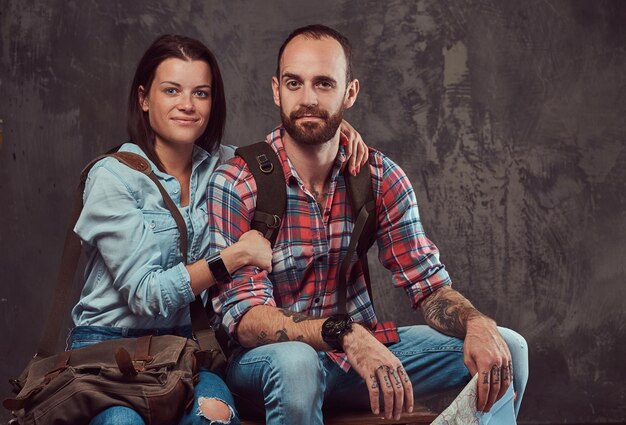 Feliz casal de turistas com mochila e mapa, abraçando enquanto está sentado em um banco em um estúdio. Isolado em um fundo cinza.