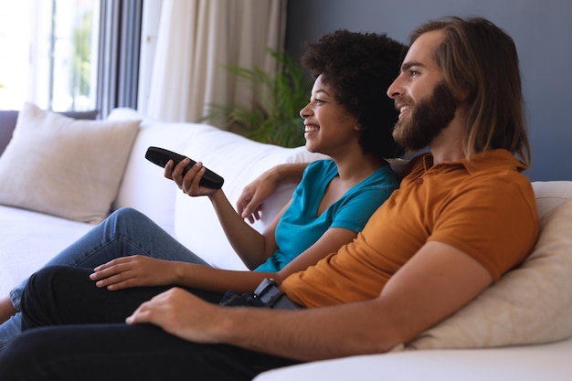 Feliz casal diversificado sentado no sofá, abraçando e assistindo tv. ficar em casa isolado durante o bloqueio de quarentena.