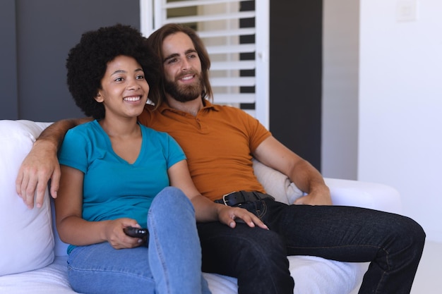 Feliz casal diversificado sentado no sofá, abraçando e assistindo tv