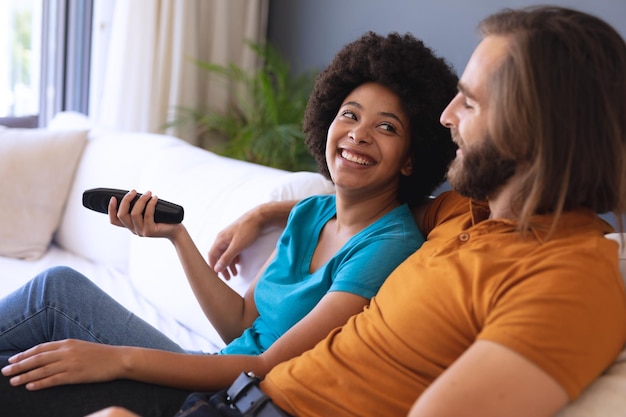 Feliz casal diversificado sentado no sofá, abraçando e assistindo tv