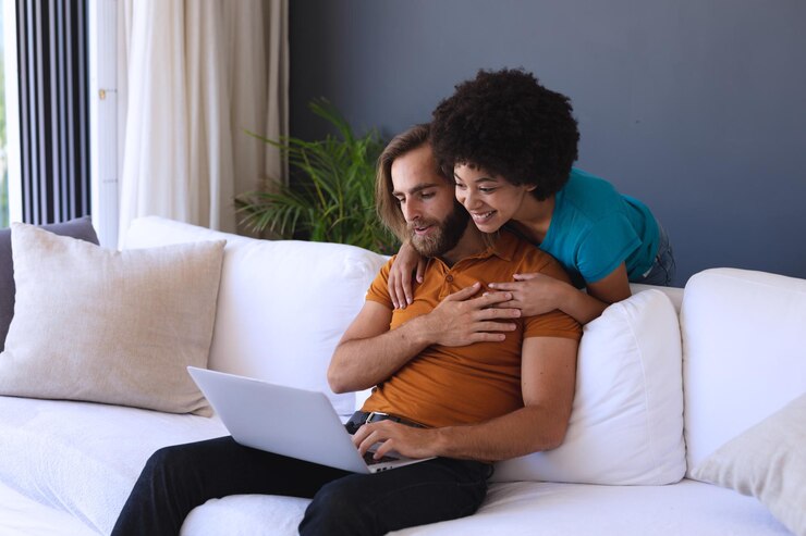 Foto feliz casal diversificado sentado no sofá, abraçando e usando laptop