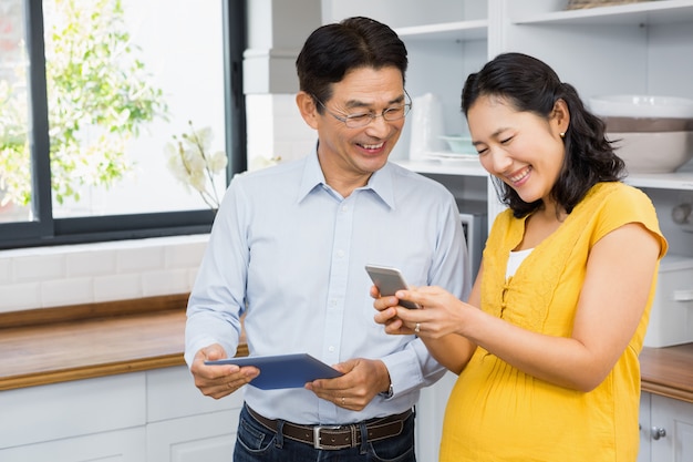 Feliz casal expectante usando tablet e smartphone na cozinha