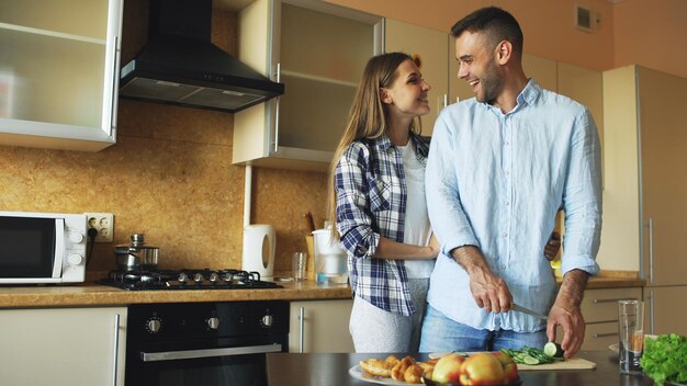 Feliz casal jovem beijando abraçando e conversando na cozinha enquanto cozinha o café da manhã em casa