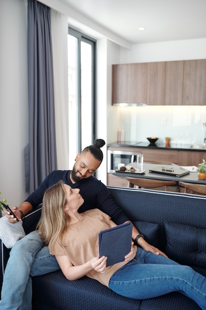 Feliz casal jovem mestiço descansando no sofá em casa e verificando as redes sociais no smartphone e no computador tablet