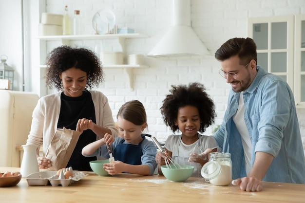 Feliz casal jovem multirracial envolvido em cozinhar pastelaria com adoráveis filhas mestiças na cozinha moderna Família diversificada sorridente preparando doces caseiros ou panquecas no café da manhã no fim de semana