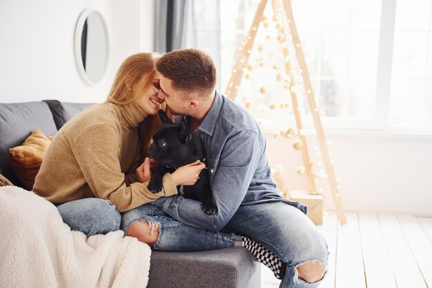 Feliz casal moderno sentado no sofá em casa com a árvore de Natal com seu cachorro fofo.