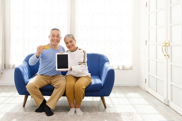 Feliz casal sênior asiático olhando para a câmera e mostrando cartão de crédito e tablet enquanto está sentado em um sofá na sala de estar Adultos idosos usando banco on-line e tecnologia Imagem com espaço de cópia