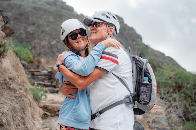 Feliz casal sênior de caminhantes com capacetes se abraçando no topo da montanha aproveitando as férias na natureza