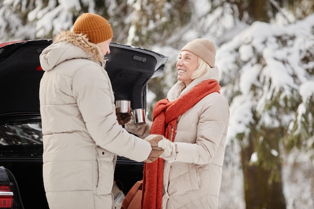 Feliz casal sênior desfrutando de uma xícara de coco quente ao ar livre na floresta de inverno e de mãos dadas