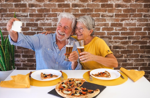 Feliz casal sênior se divertindo junto com pizza e cerveja Mulher idosa e homem rindo sentado na mesa de casa usando telefone para uma selfie