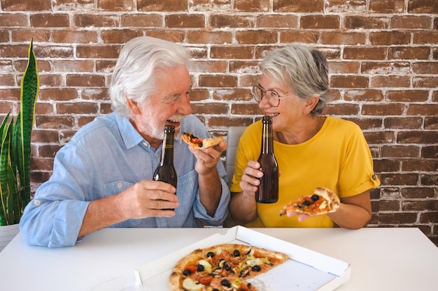 Feliz casal sênior se divertindo juntos comendo uma pizza Sentado na mesa de casa com cerveja e pizza Celebrando férias em companhia