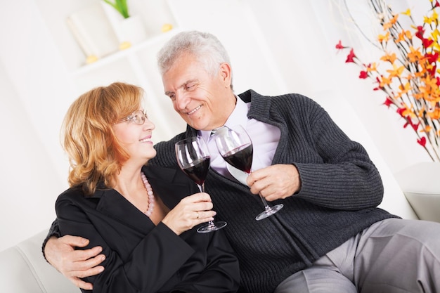 Feliz casal sênior sentado abraçado em casa, sorrindo e bebendo vinho tinto.
