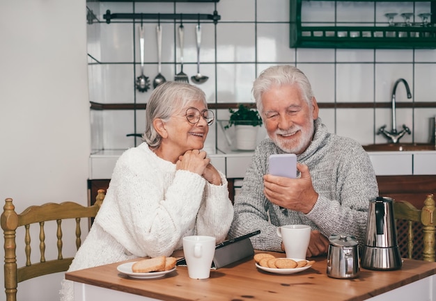 Feliz casal sênior tomando café da manhã juntos à mesa em casa olhando para o celular