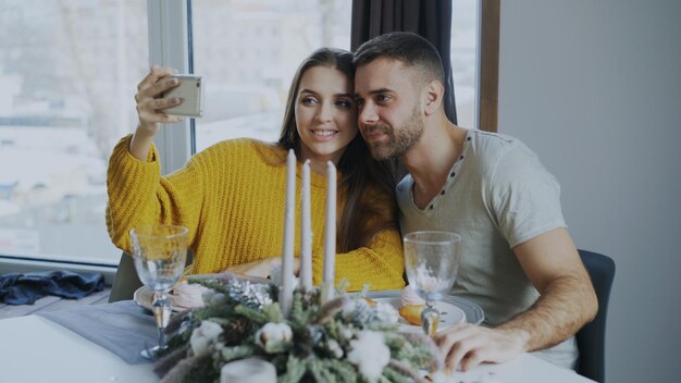 Feliz casal sorridente almoçando e tirando retrato selfie com smartphone no café dentro de casa