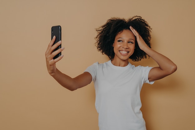 Feliz charmoso feminino de pele escura de 20 anos em camiseta branca, fazendo selfie de foto no smartphone moderno e sorrindo alegremente, isolado sobre a parede bege. Mulher africana jovem positiva tirando foto no celular