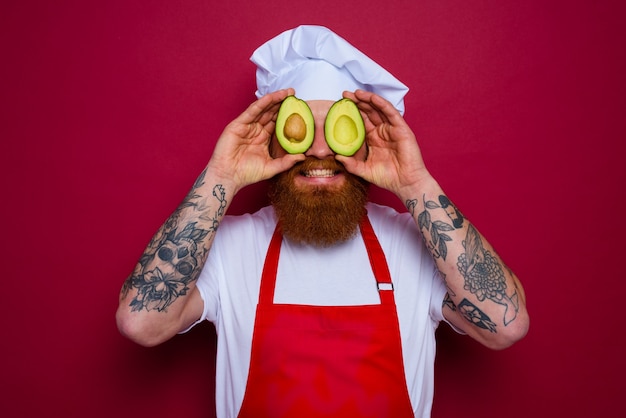 Feliz chef con barba y delantal rojo sostiene un aguacate